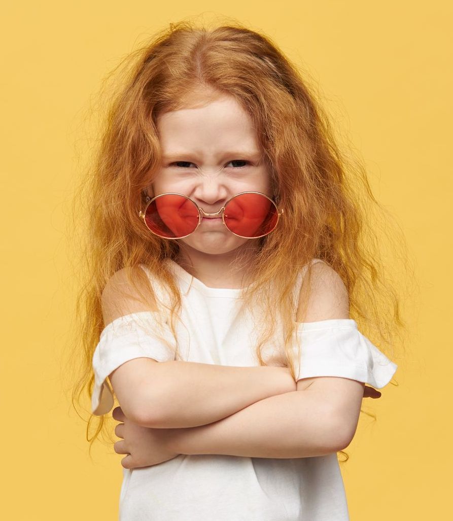 Ragazza con i capelli rossi e occhiali da sole, braccia incrociate, accigliata su sfondo giallo.
