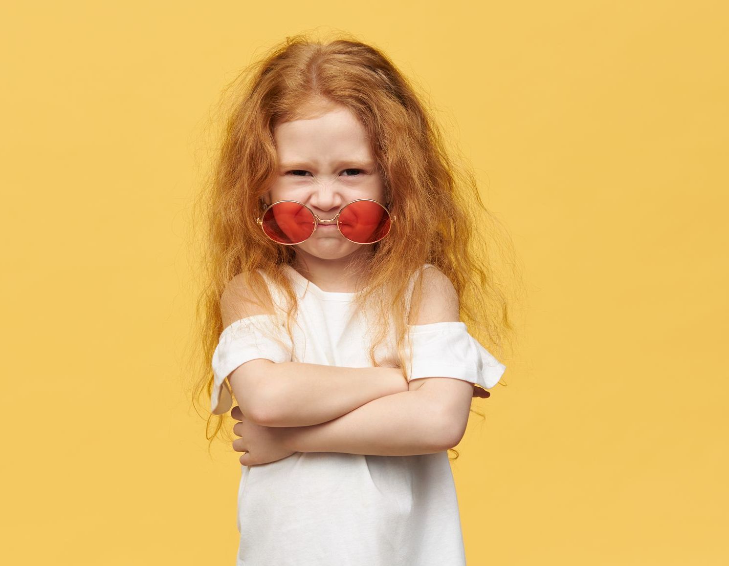 Bambino dai capelli rossi con occhiali da sole a forma di cuore, braccia incrociate e sguardo accigliato, su sfondo giallo.