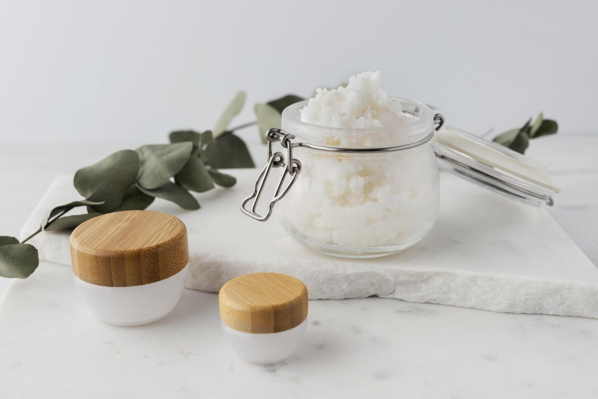 Barattolo di burro di karité, due piccoli contenitori con coperchi di legno ed eucalipto su una superficie di marmo bianco.