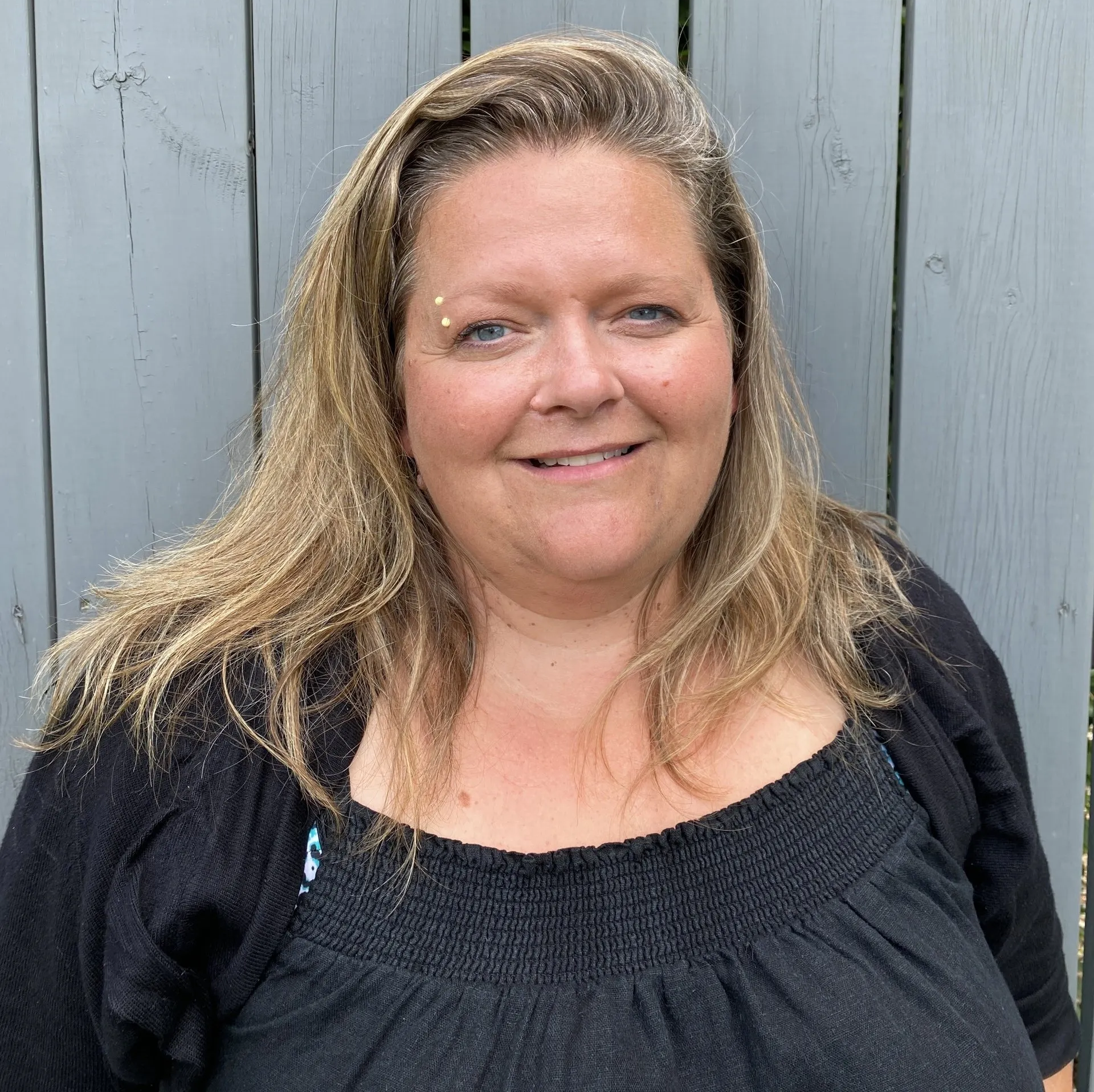 A woman in a black shirt is smiling in front of a wooden fence.