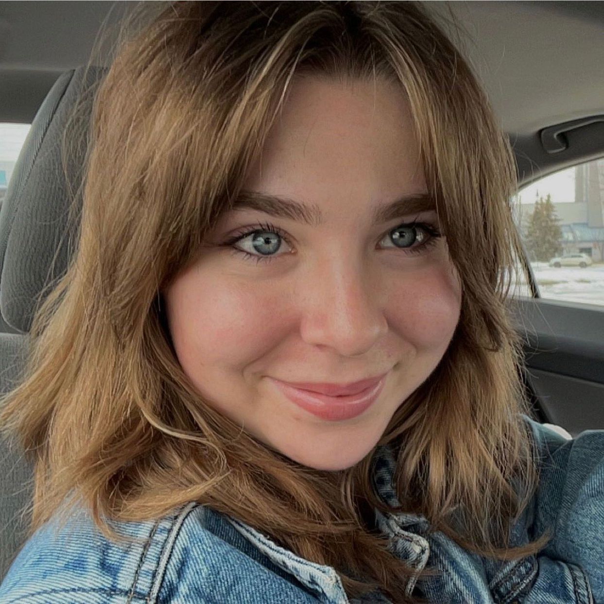 A woman is smiling while sitting in a car wearing a denim jacket.