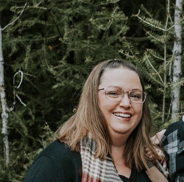 A woman wearing glasses and a scarf is smiling in front of trees.