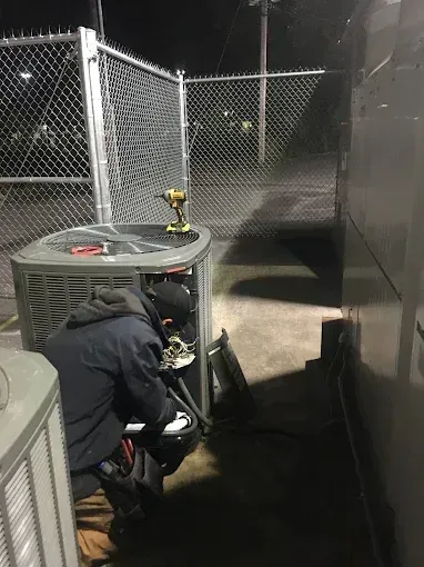 A technician in a dark jacket repairs an outdoor HVAC unit at night near a chain-link fence.