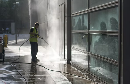 Worker pressure-washing a glass building facade on a wet sidewalk in bright sunlight