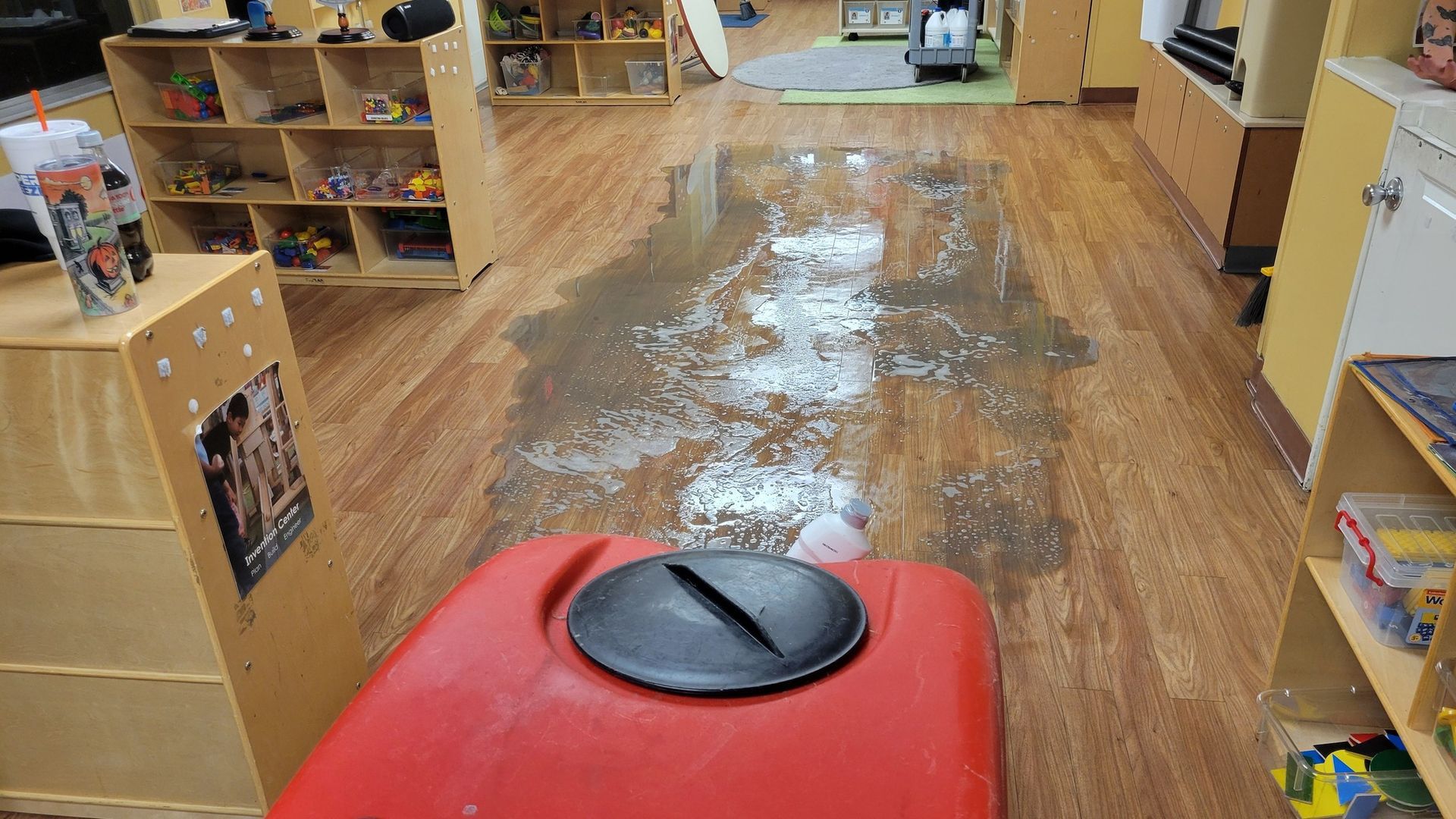 A flooded daycare room with water on the wooden floor and a red cleaning machine.