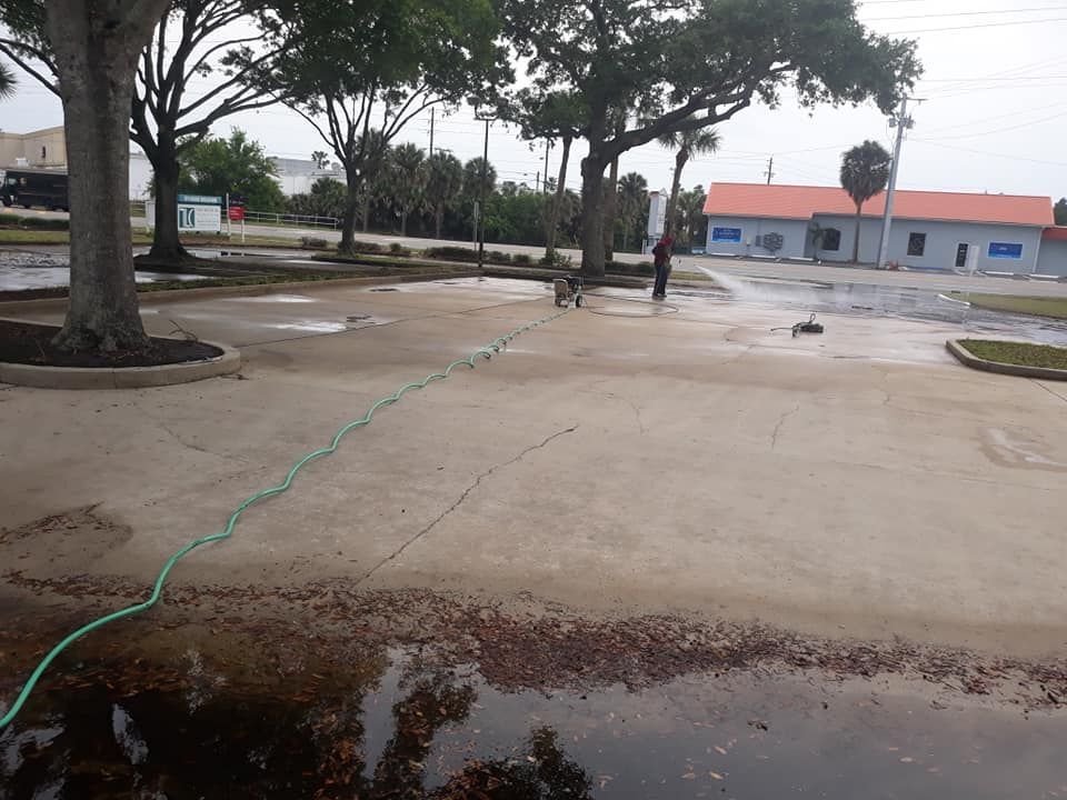 Man washing a concrete parking lot with water hose and tools near a building and trees.