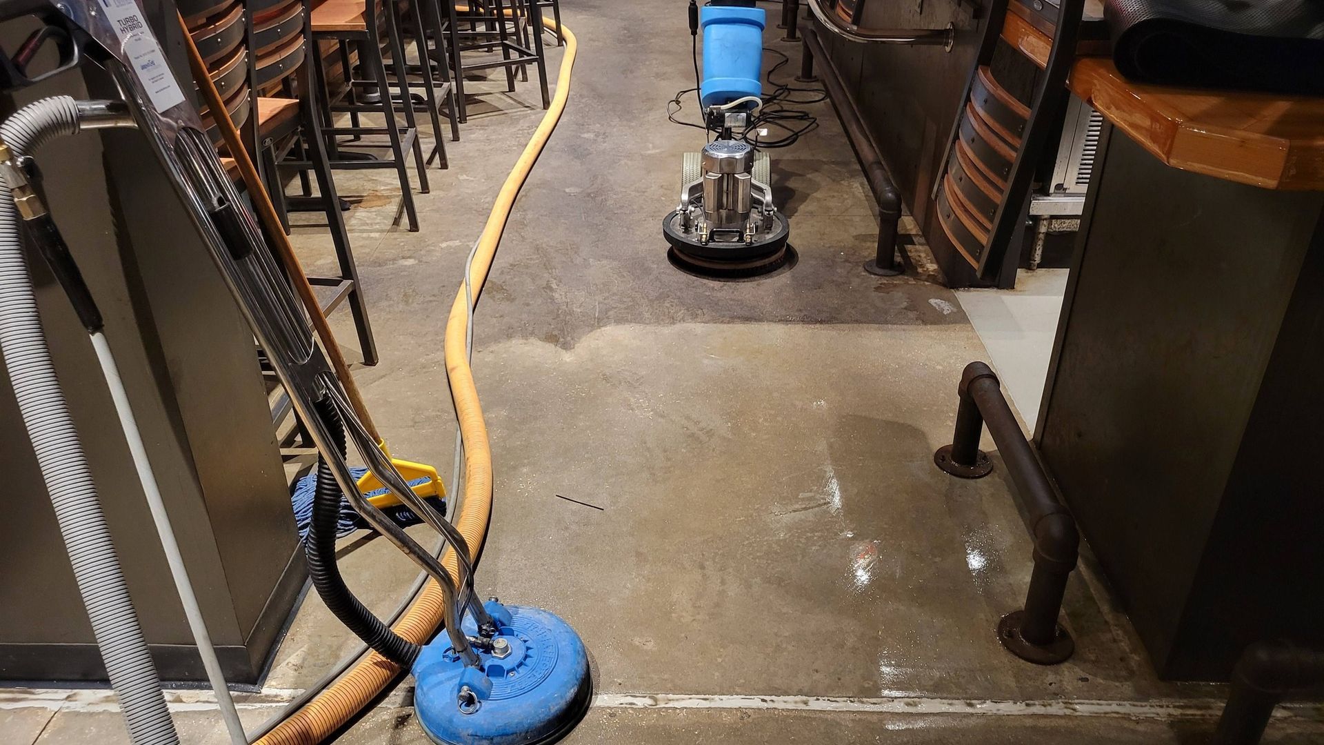 Floor cleaning machine in use, wet floor, bar setting. Blue equipment, yellow hose.