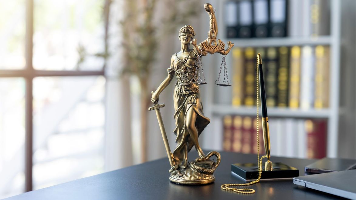 Gold Lady Justice statue on a desk beside a gavel and scales, with bookshelves in the background