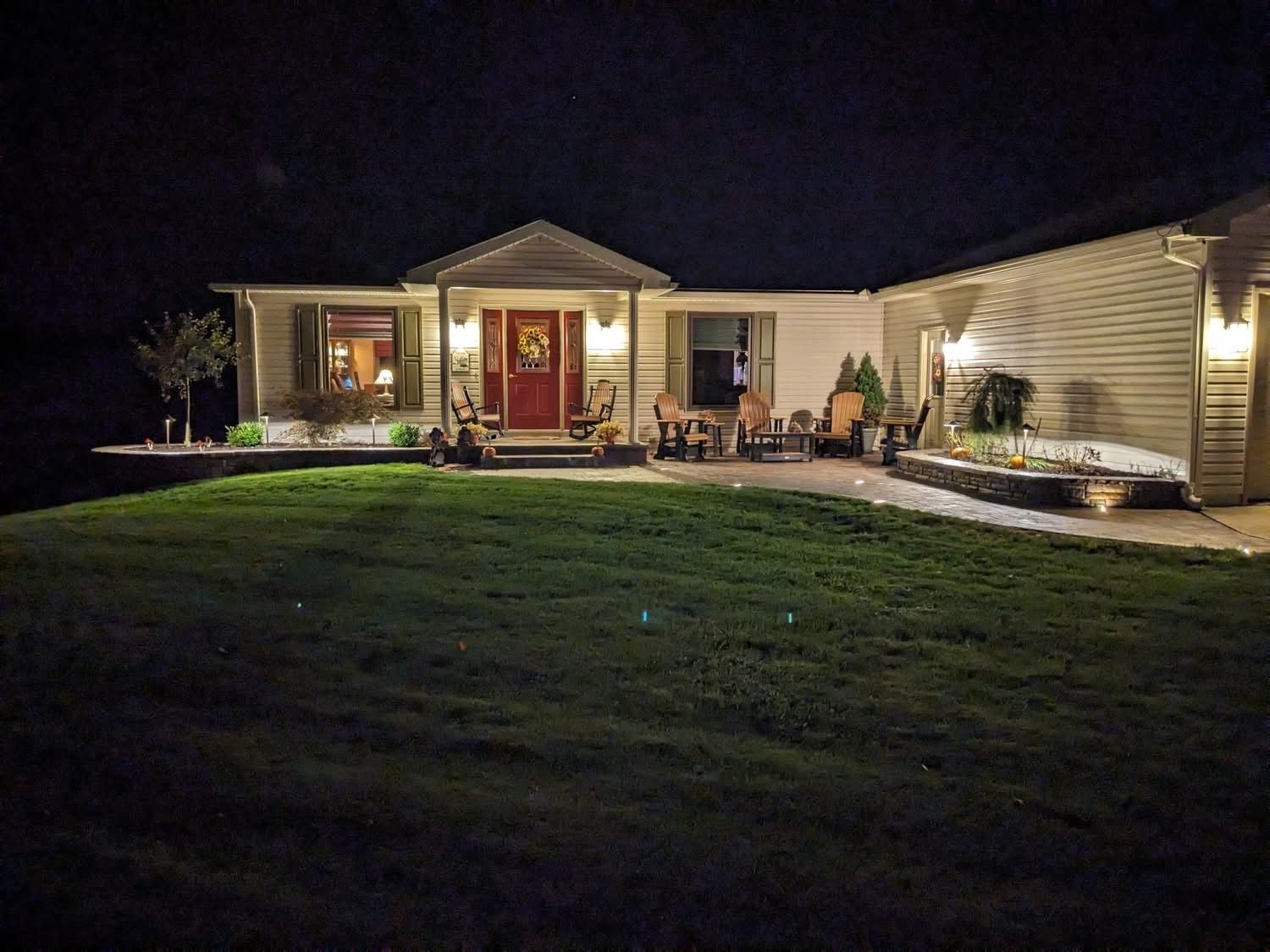 House exterior at night, with illuminated porch and red front door, lawn, and landscaping.