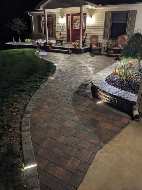 Brick pathway leads to a house with porch, lit by outdoor lights at night.