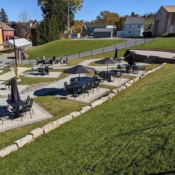 Outdoor seating area on a grassy hill; tables with umbrellas, stone retaining walls, buildings in the background.