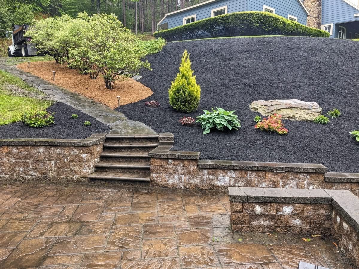 Stone steps and patio lead to a landscaped hillside with dark mulch and various green plants.