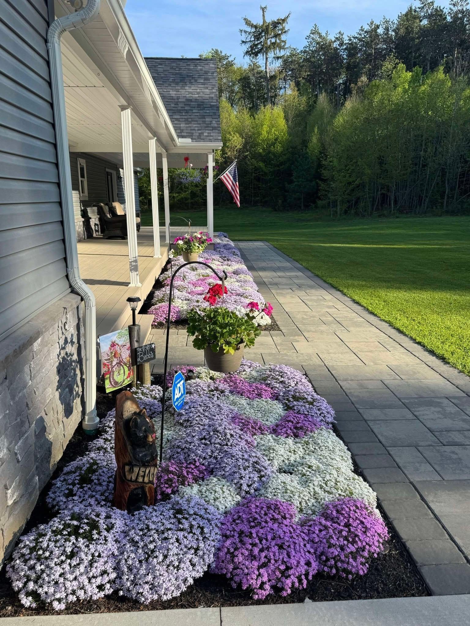 A house with a porch and flower beds. Purple, pink, and white flowers line the walkway. An American flag hangs.