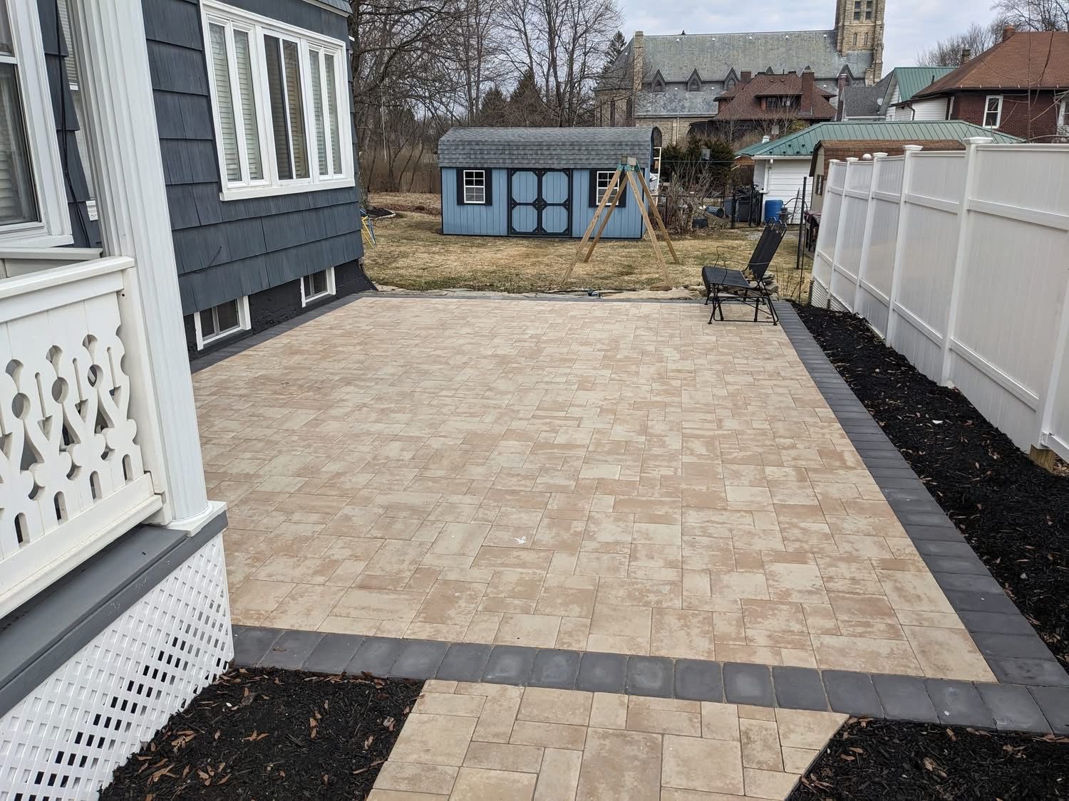Paved patio with tan pavers, dark border, and white fence. Shed and building in the background.