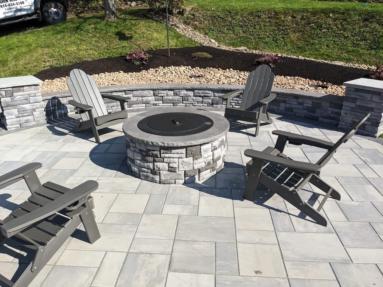 Stone fire pit surrounded by four Adirondack chairs on a paved patio.