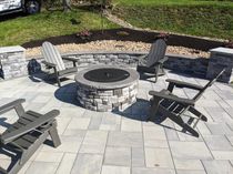 Stone fire pit surrounded by four Adirondack chairs on a paved patio.