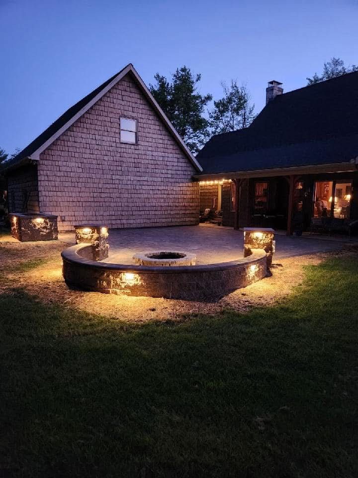 Nighttime patio with fire pit, stone walls, and warm lighting near a house.