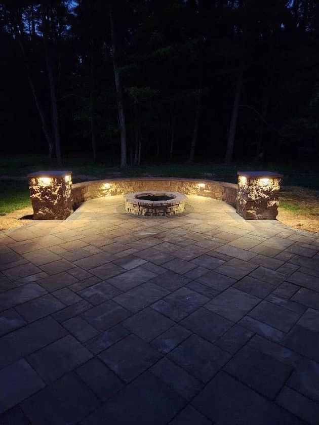 Nighttime shot of a patio with a fire pit, stone wall, and illuminated pillars. Dark trees in the background.