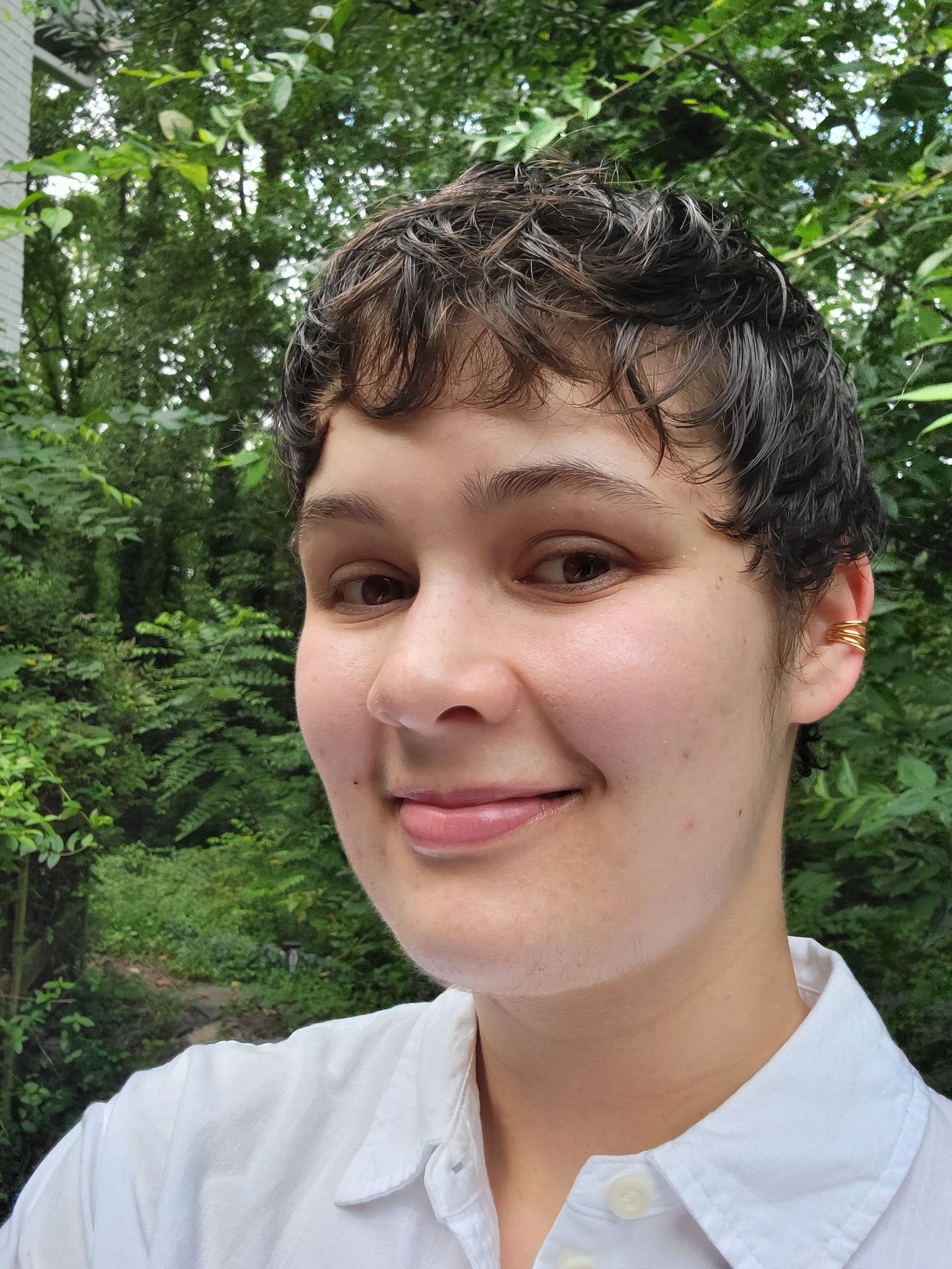 Smiling person with short curly hair in a white shirt, standing outdoors among green trees.