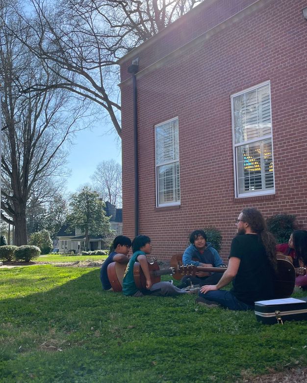 People sitting on grass beside a brick building in a sunny park-like setting
