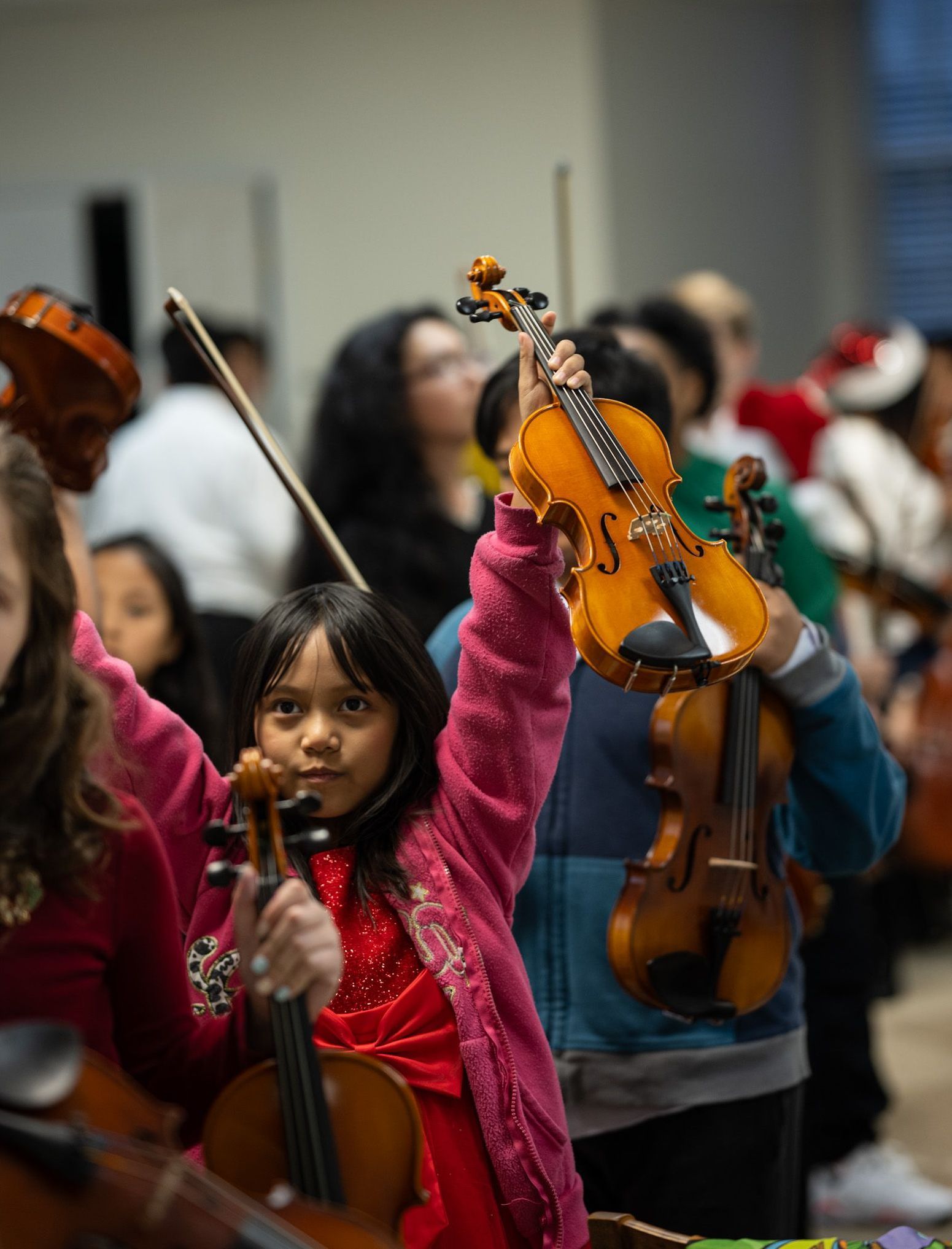 Children in a music class holding violins, one raising an orange violin in front of a blurred audience