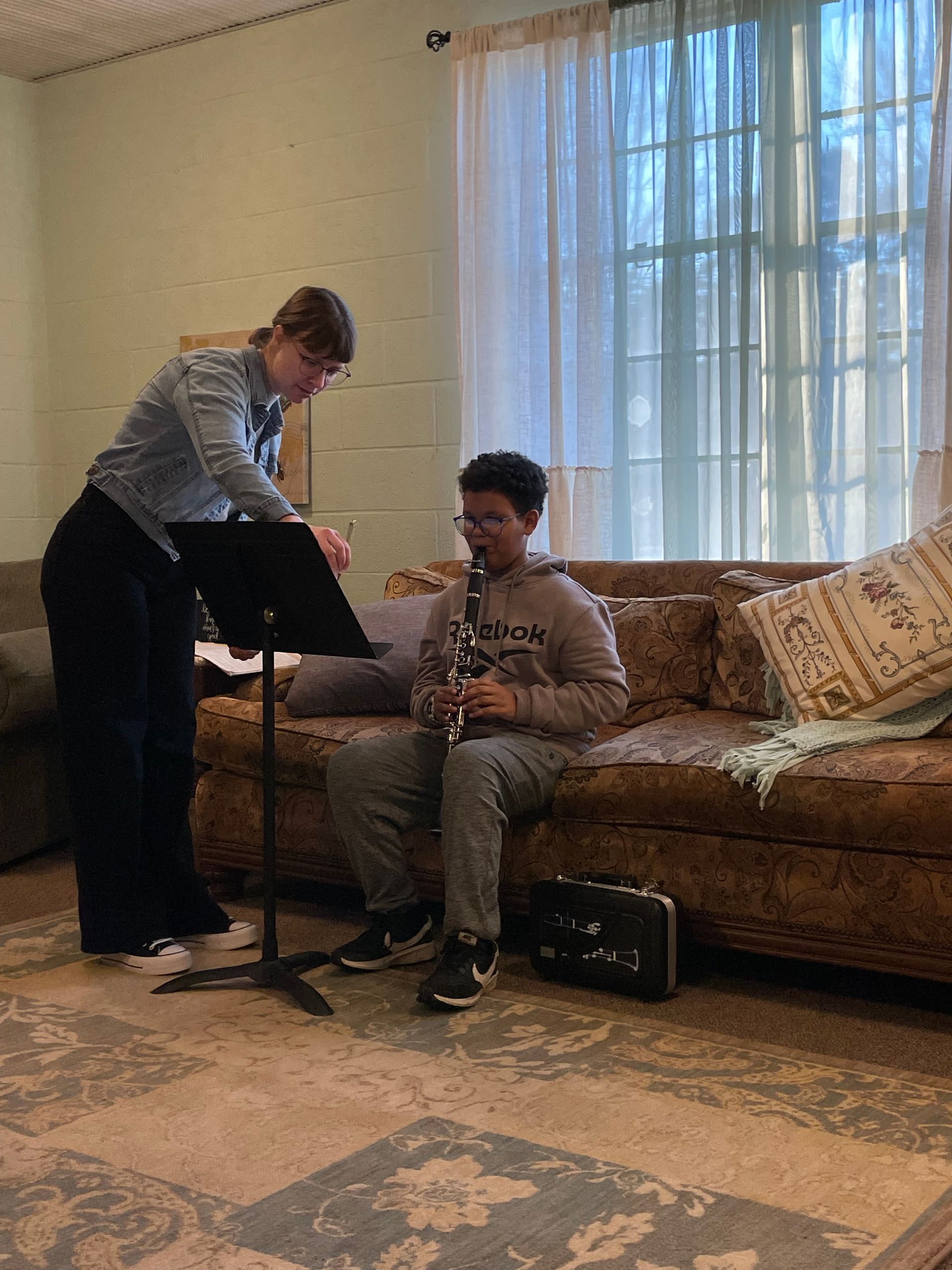 Two people rehearsing music in a living room, one adjusting a microphone stand as the other sits on a couch holding a guitar.