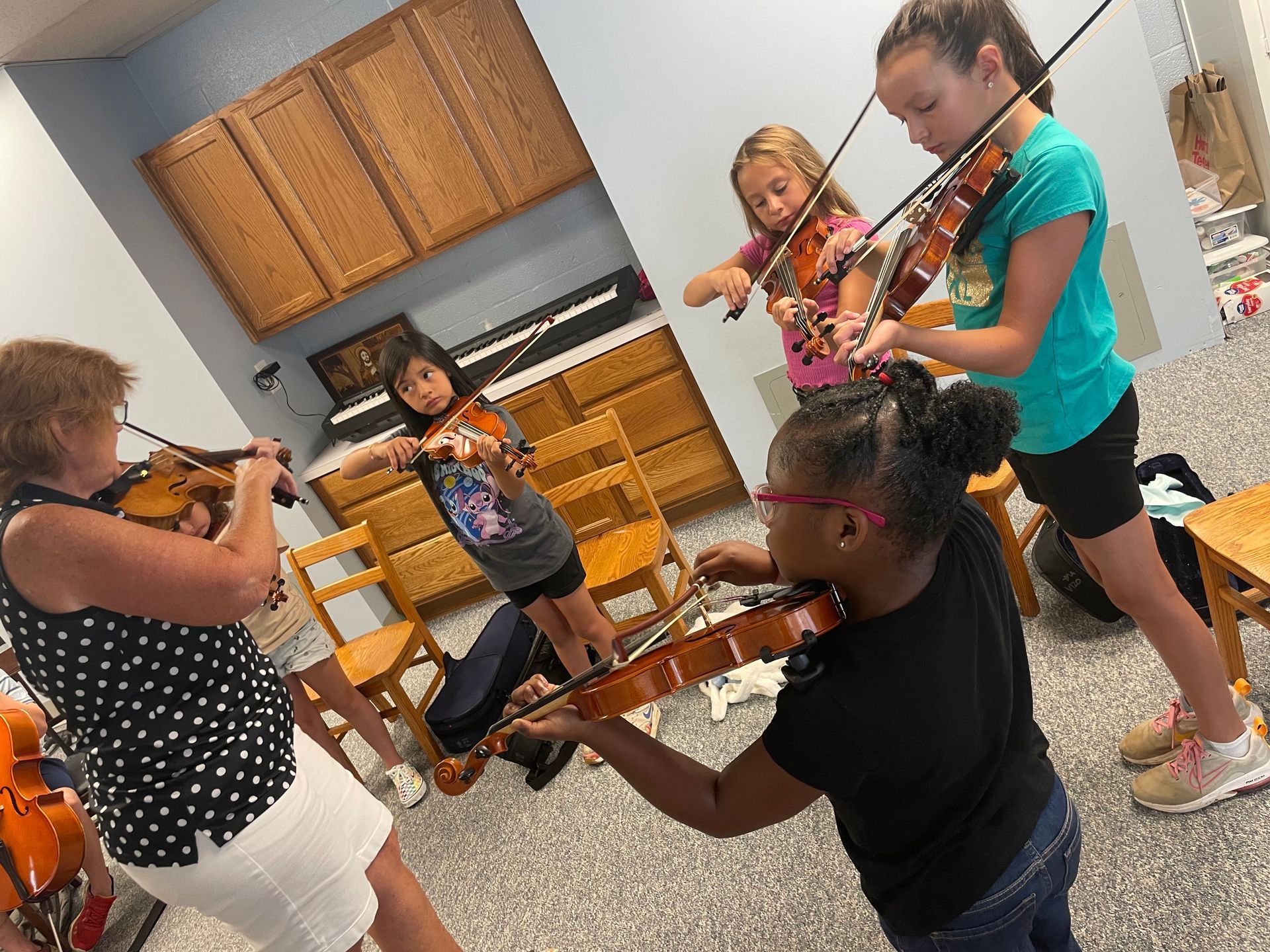 Children playing violins together in a bright kitchen, with sheet music and wicker drawers in the background
