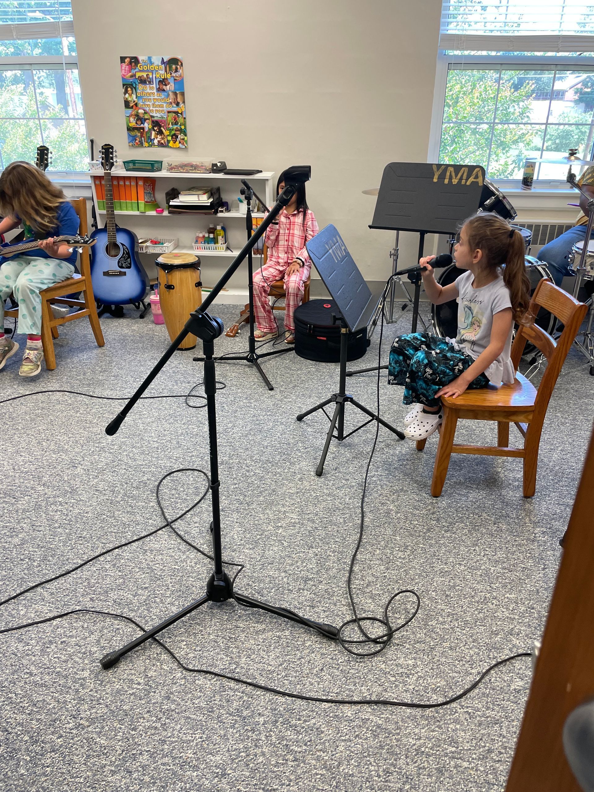 Children sitting with guitars in a bright room, with music stands and a microphone in the foreground.