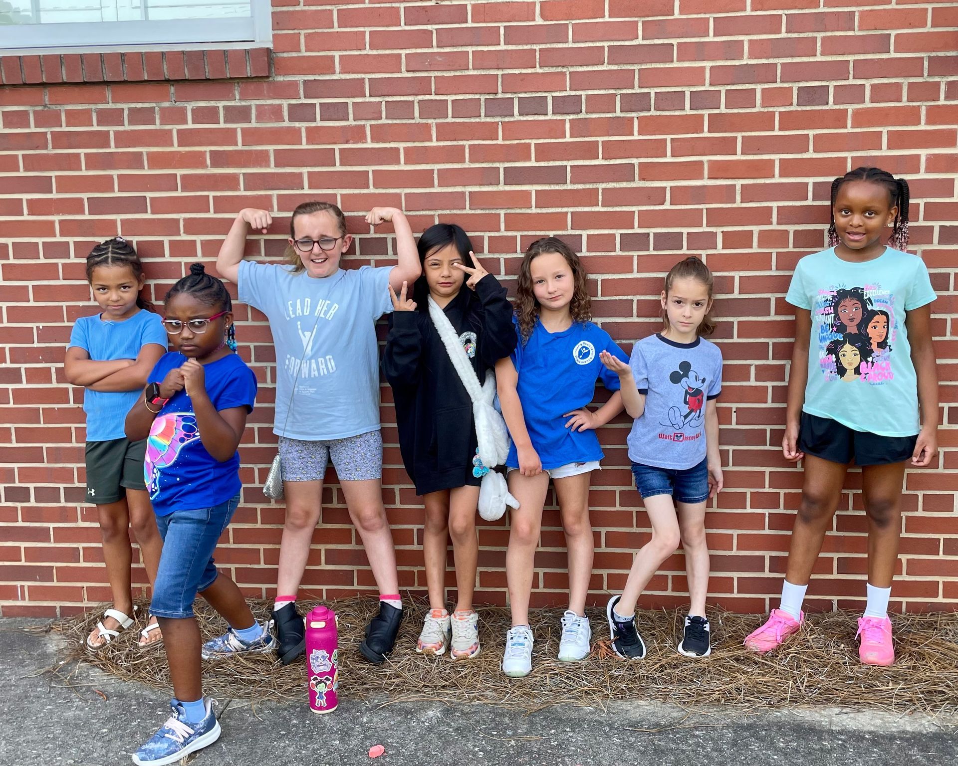 Six children posing with arms raised in front of a brick wall, wearing colorful T-shirts and shorts.