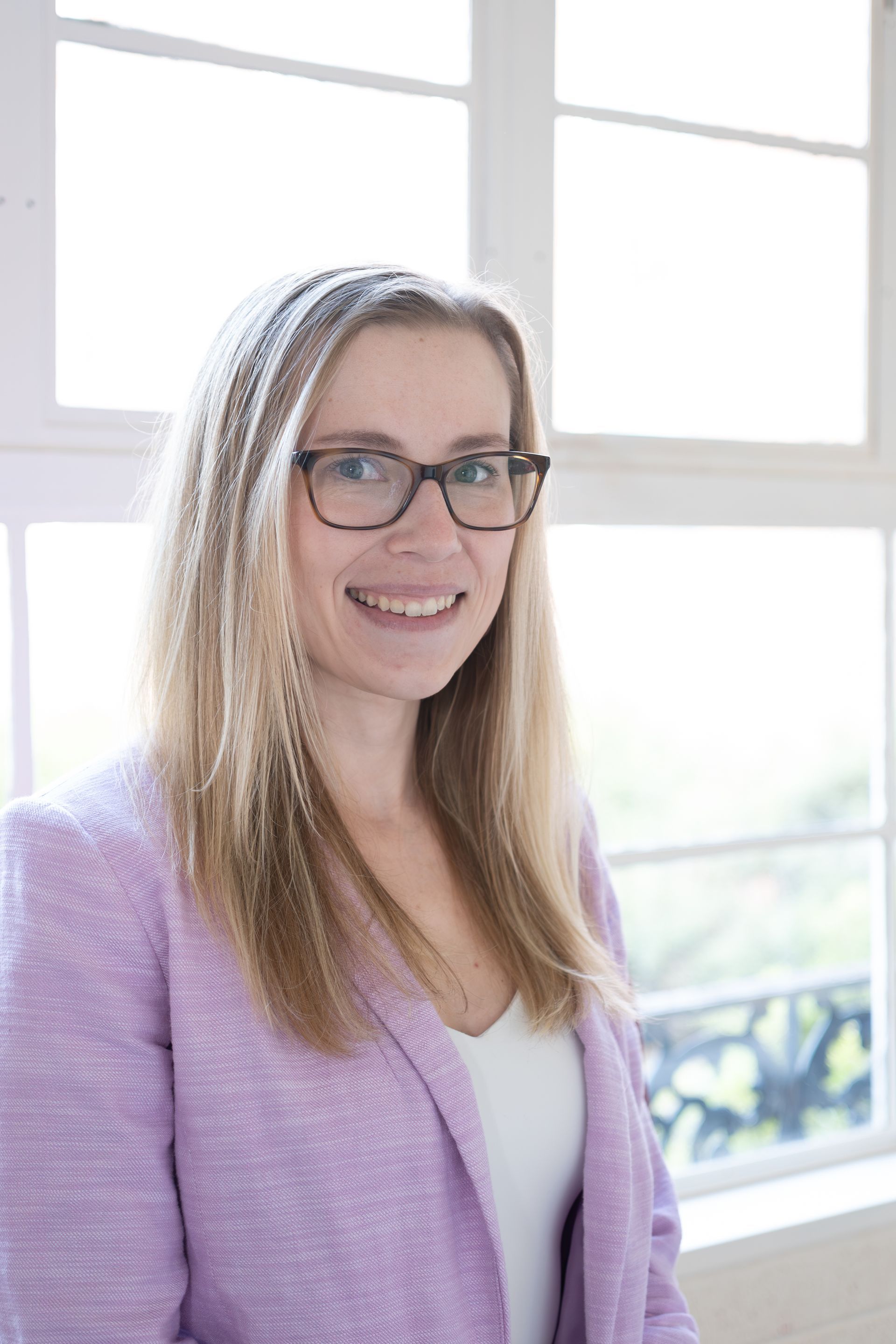Smiling person with glasses in a lavender cardigan standing by a bright window