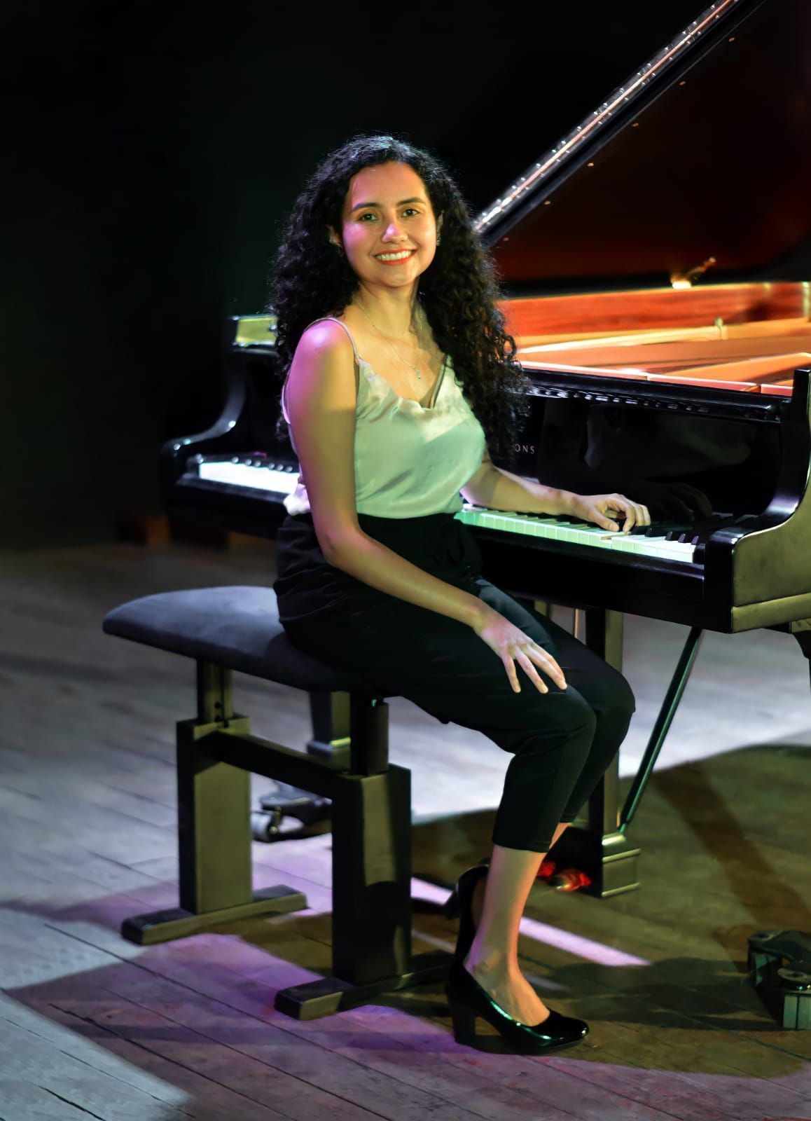 Woman seated on a piano bench beside a grand piano, smiling in a dimly lit performance setting