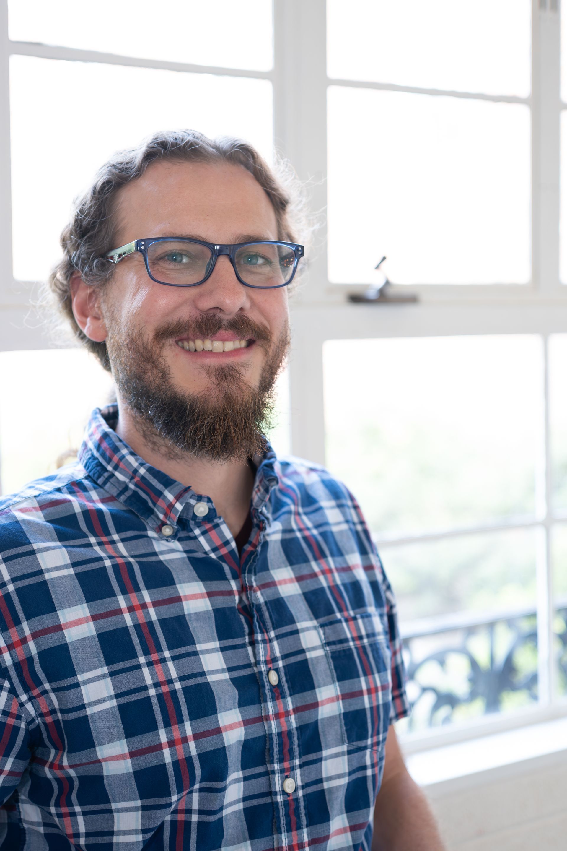 Smiling person in glasses and a blue plaid shirt standing by a bright window