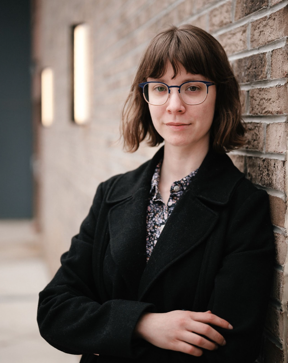 Person with glasses and black coat leaning against a brick wall in a hallway.