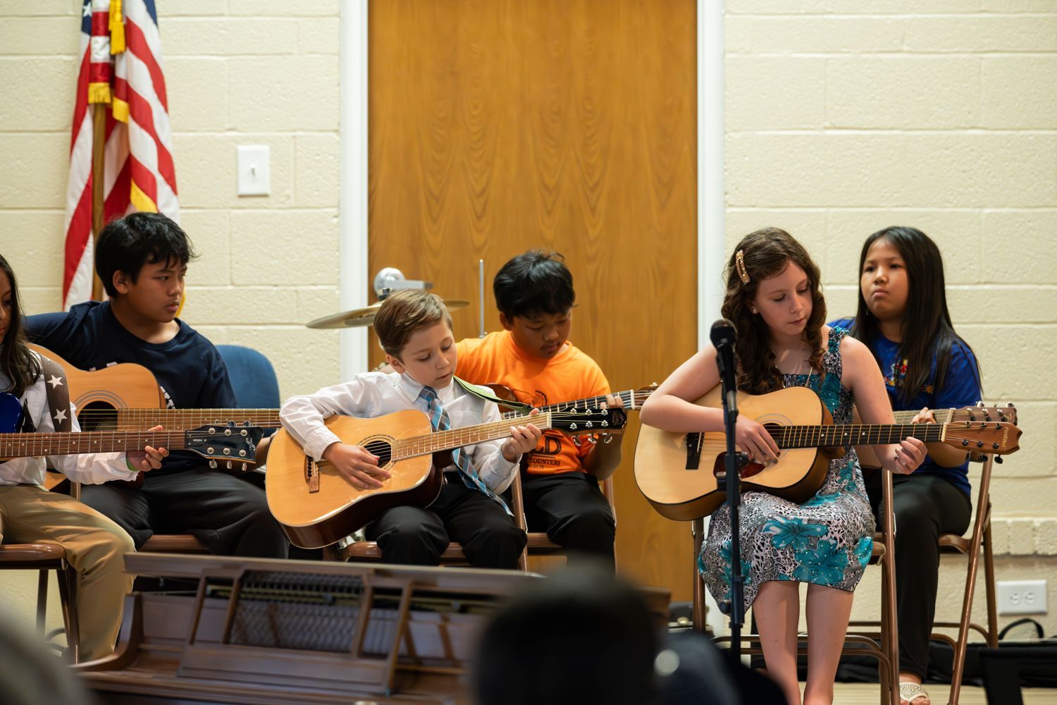 Children seated on stage, playing acoustic guitars during a classroom performance