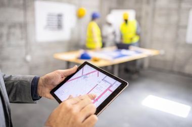 Hands using a tablet with a site plan in front of blurred construction workers at a table