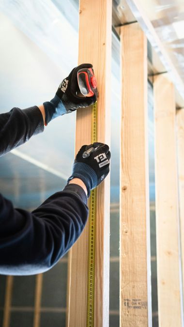 Gloved hands measuring a vertical wooden stud with a tape measure during construction