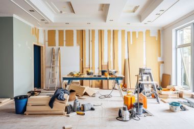 Renovation in progress in a bright room, with tools, ladders, paint supplies, and exposed wall framing.