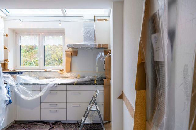 Kitchen under renovation, with covered cabinets, ladder, and plastic sheeting by a bright window.
