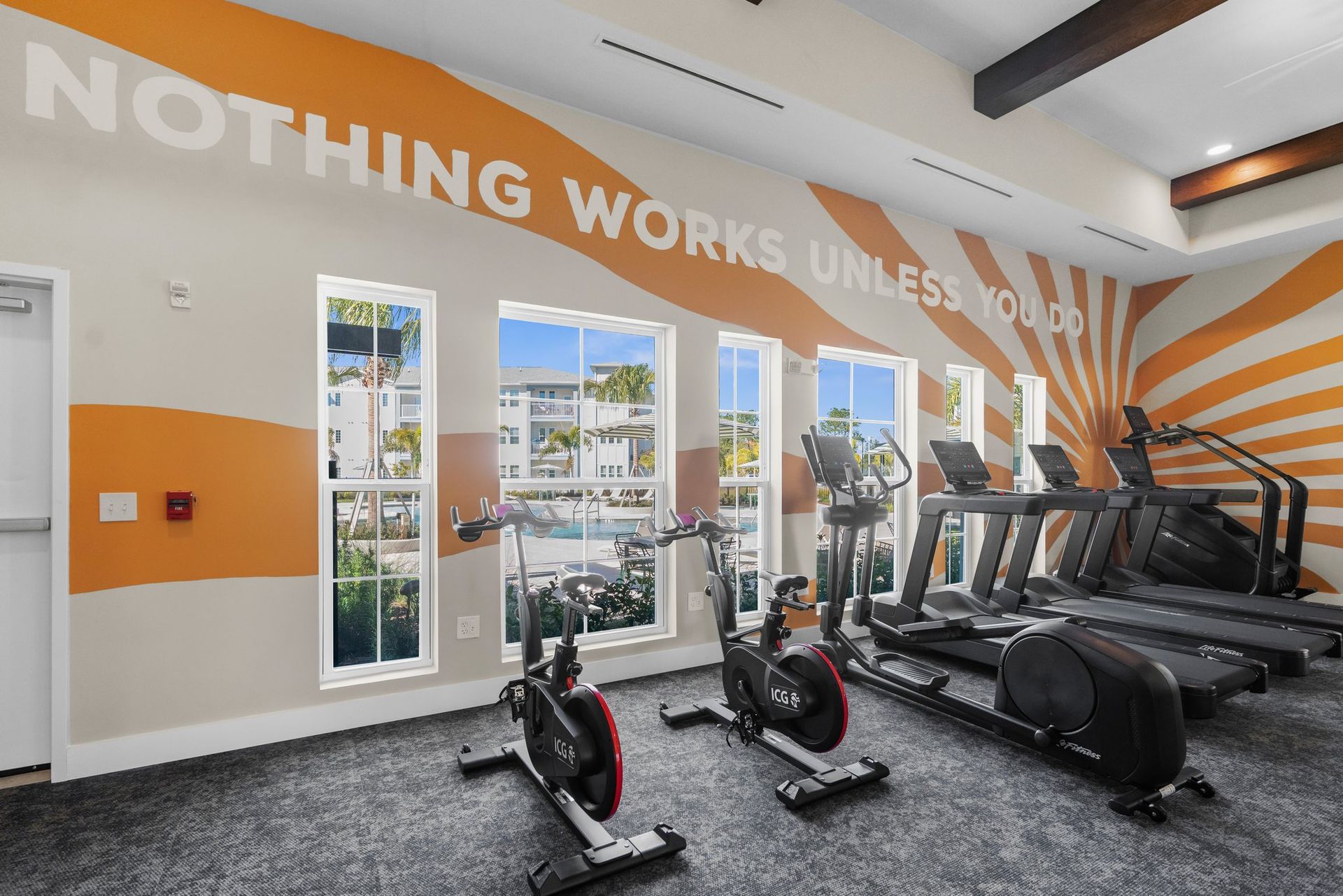 Gym with exercise machines, windows, and motivational wall art in orange and white.