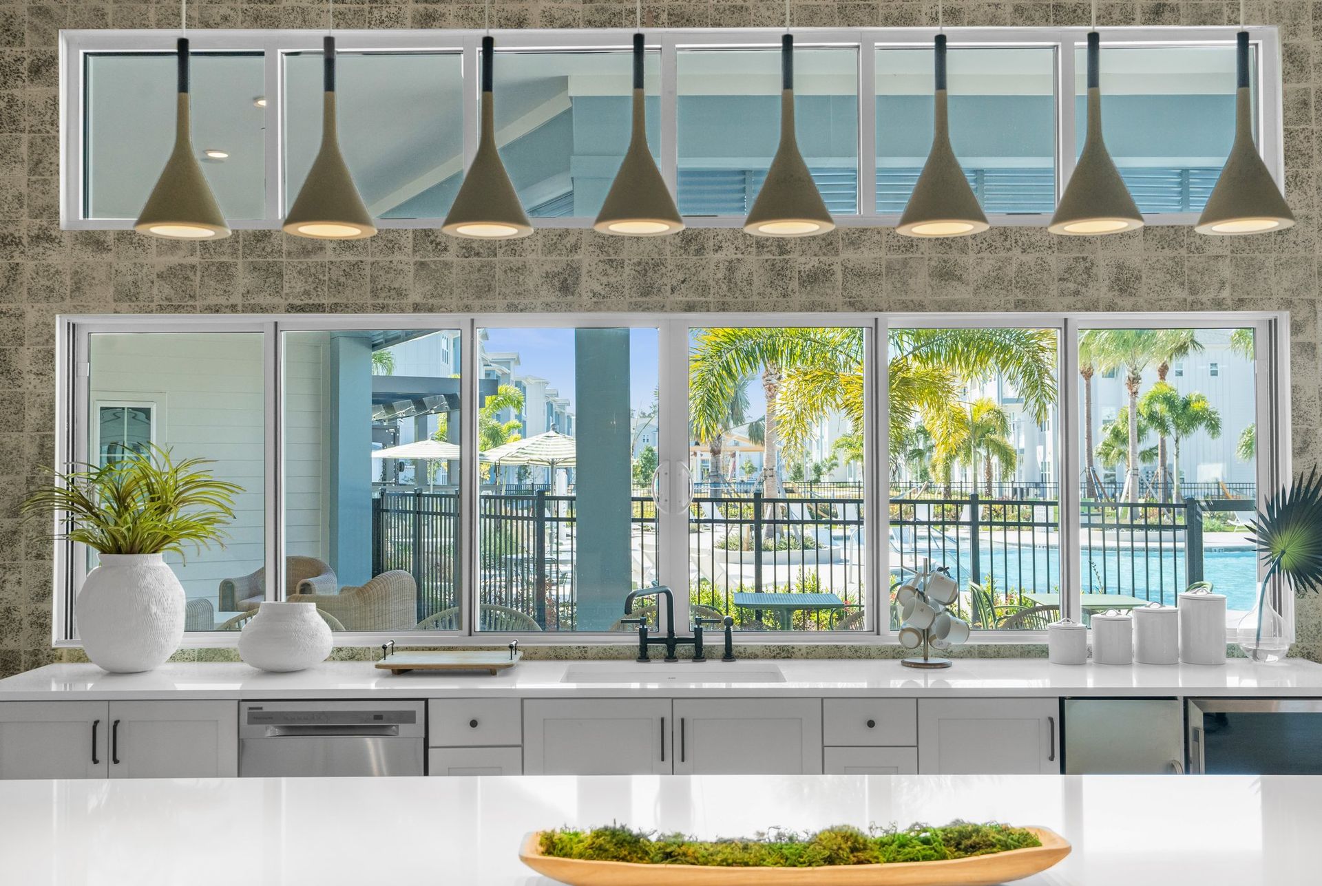 Kitchen with long windows overlooking a pool, light fixtures, white counters, and greenery.