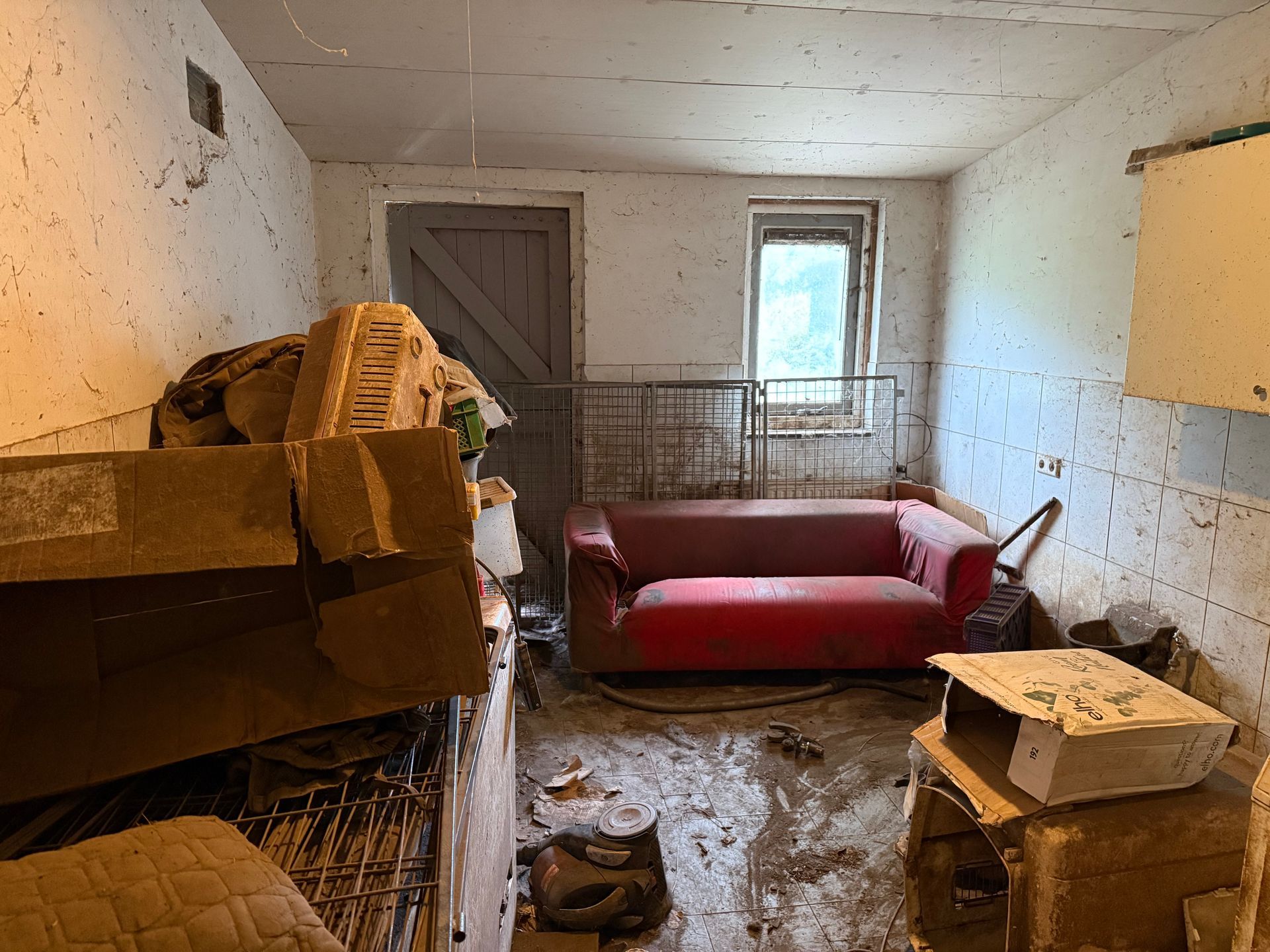 Een rommelige, vieze kamer met een rode bank, kartonnen dozen en een raam.