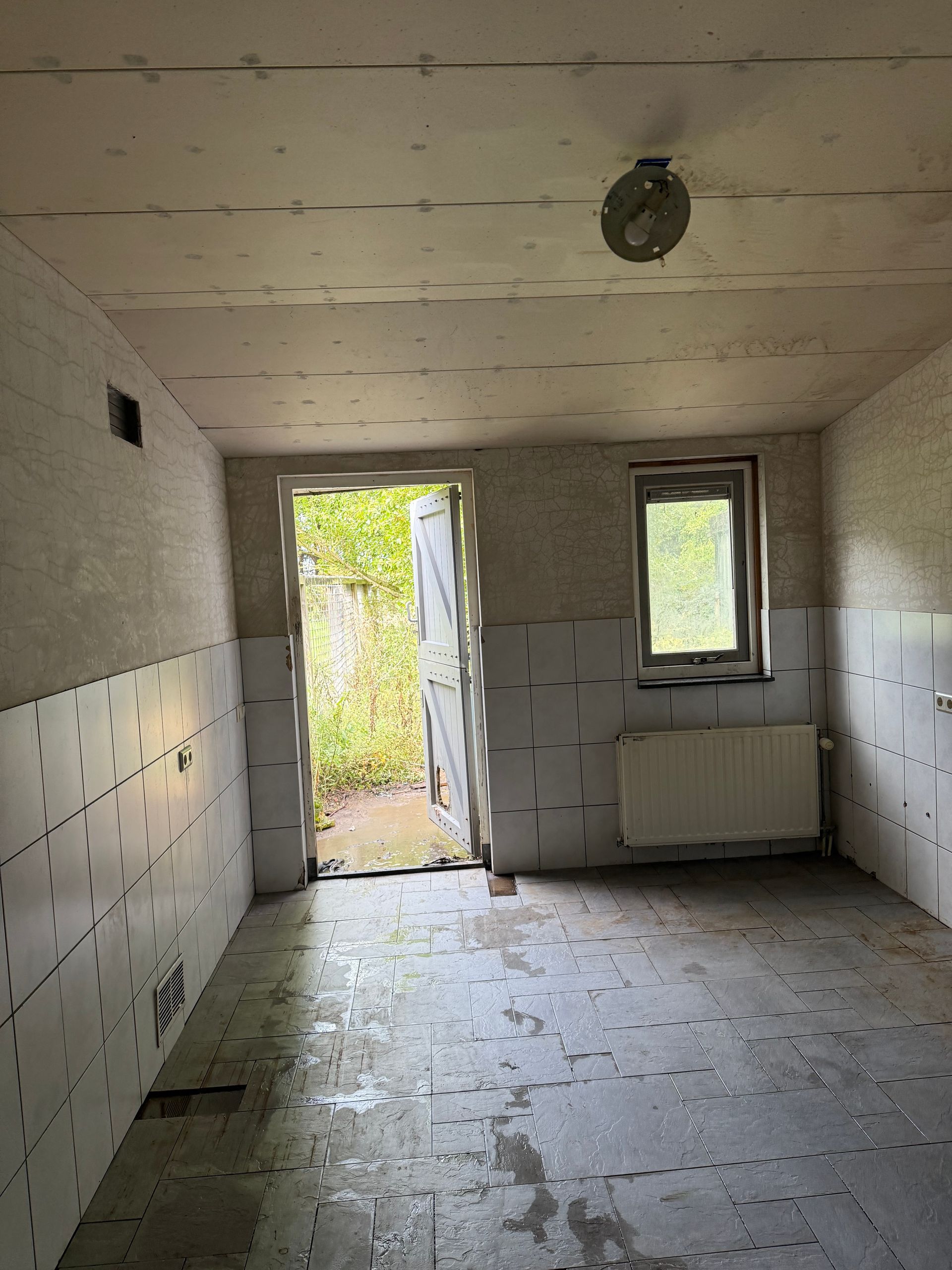 Kamerinterieur, onafgewerkte constructie. Kale muren, zichtbaar plafond, open deur naar buiten, klein raam, radiator.