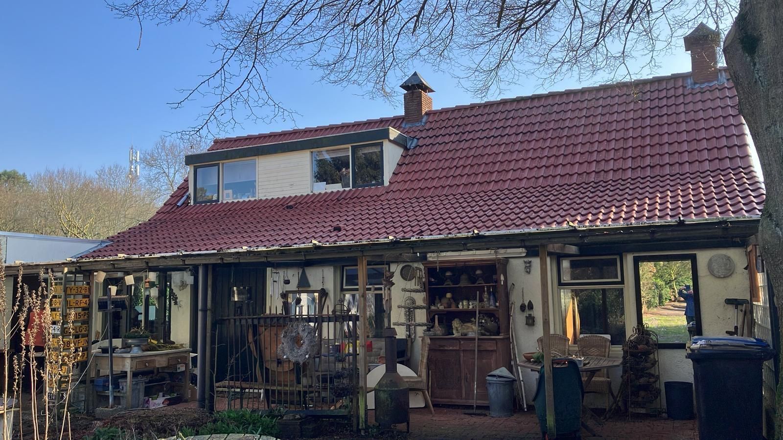 Een huis met een rood pannendak, een overdekte veranda en een rommelige tuin tegen een blauwe hemel.