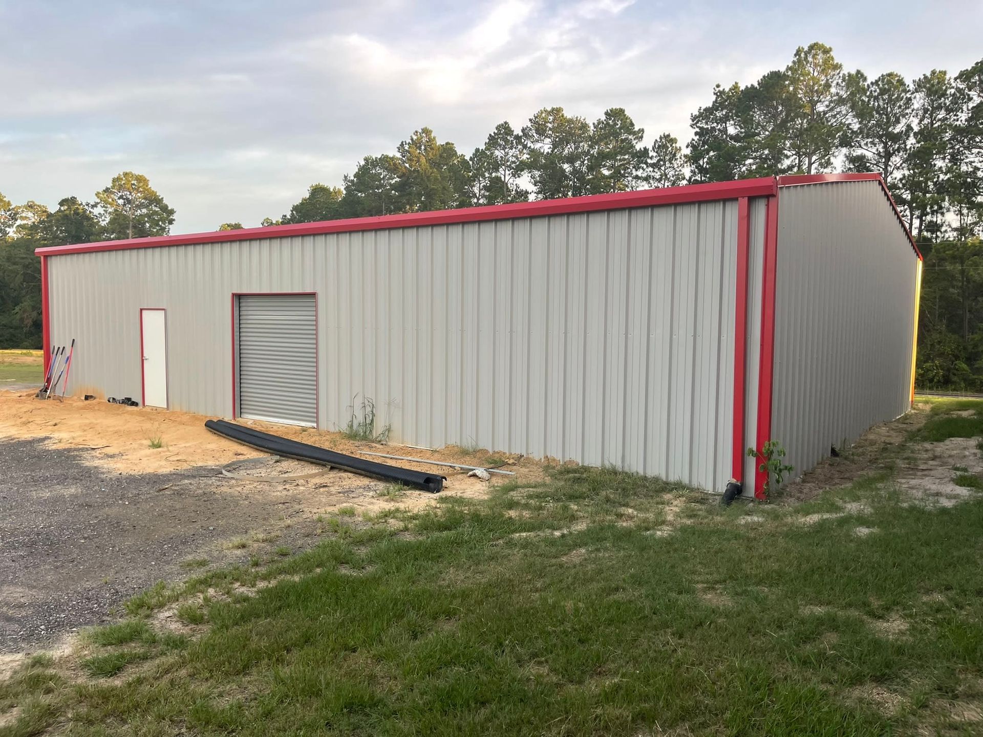 Metal building with red trim, a door, and a garage door, set on a grassy and gravel area under a cloudy sky