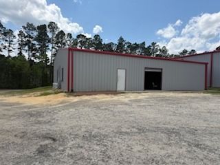 Gray metal industrial building with red trim, white door, and open garage door on a gravel lot; trees in the background.