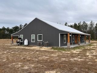 Gray metal-sided house with white roof and covered porch in a field. Includes propane tank and grill.