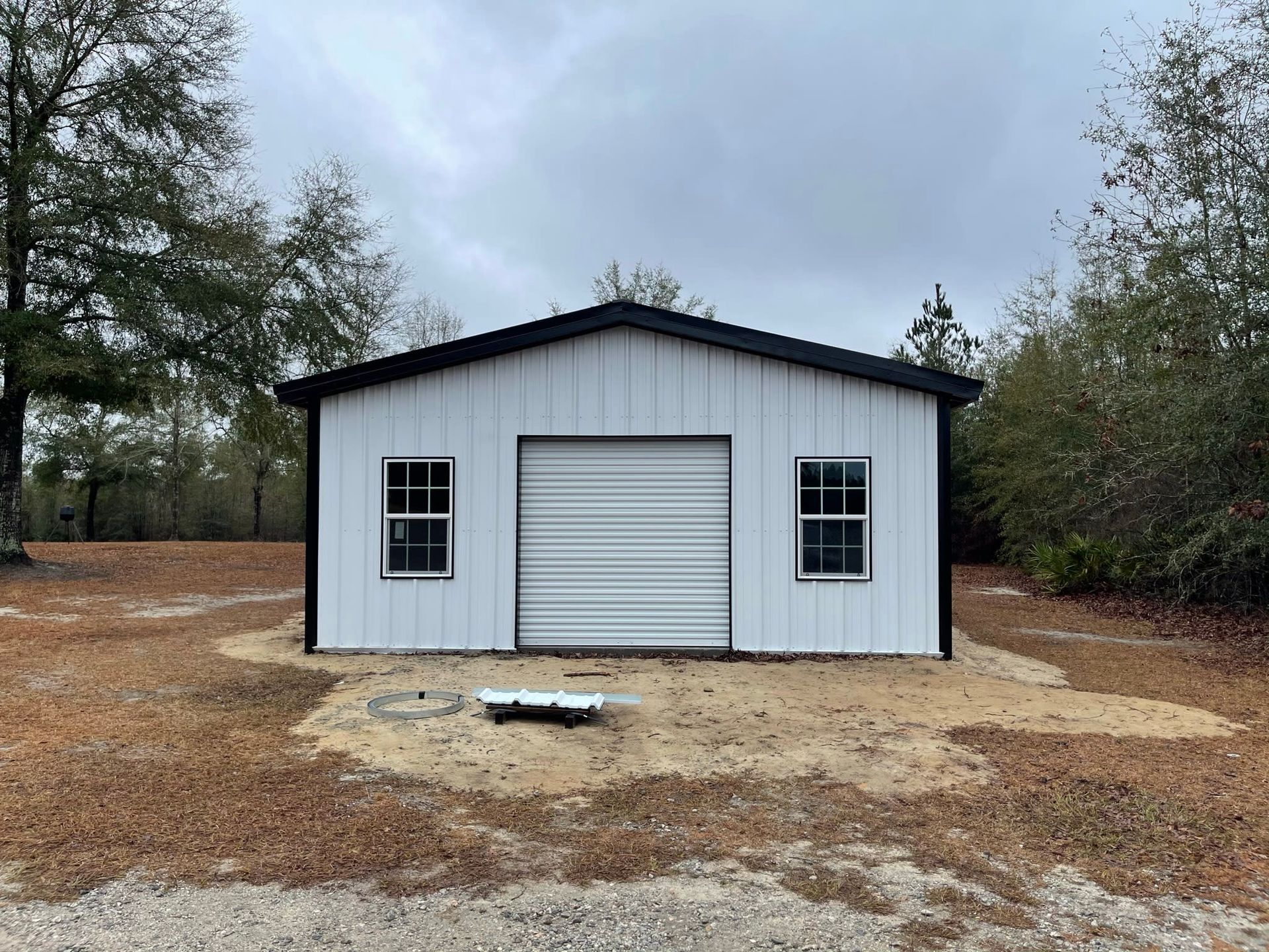 White metal garage with black trim, roll-up door, and windows in a wooded area