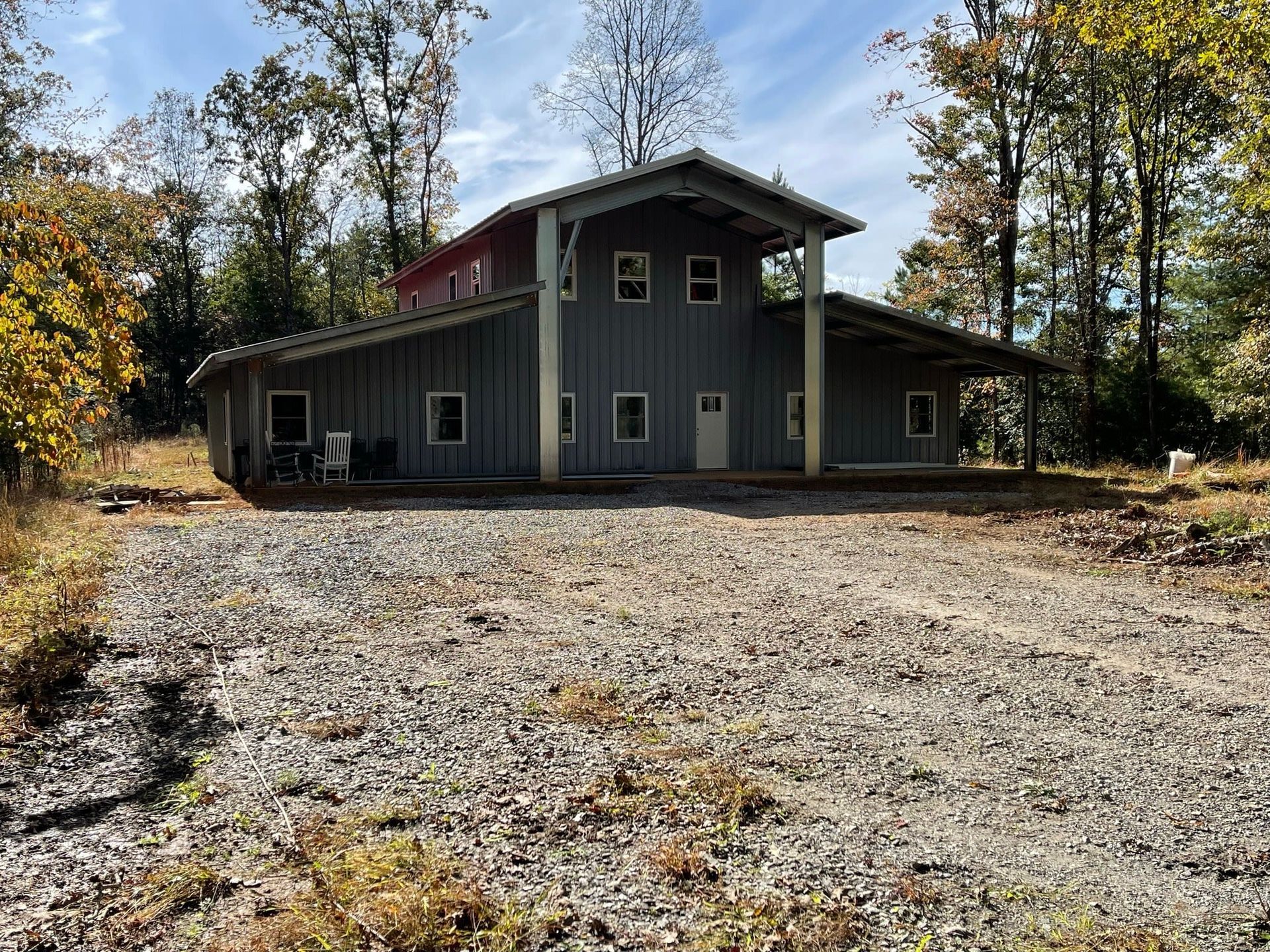 Gray house with gabled roof and covered porch, gravel driveway, surrounded by trees.