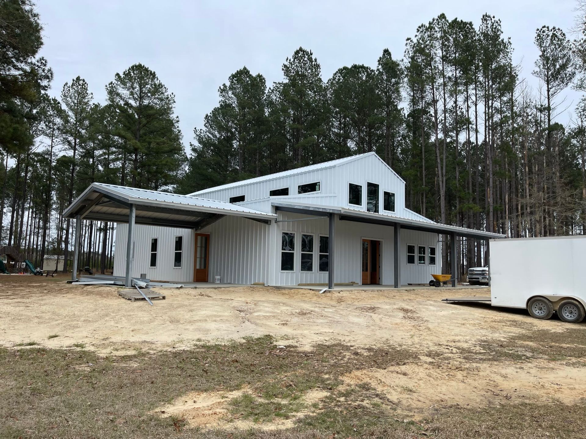 White barn-style building under construction with metal roof and covered porch, set in a clearing near trees