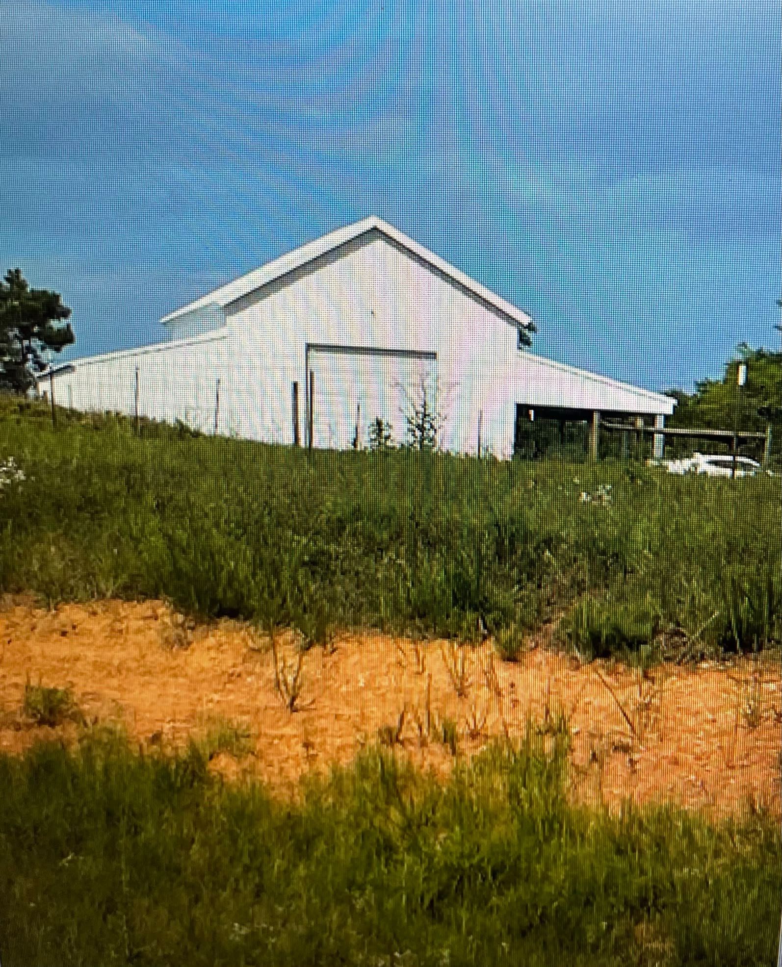 White barn on a grassy hillside under a blue sky, with a fenced area and dirt in the foreground.